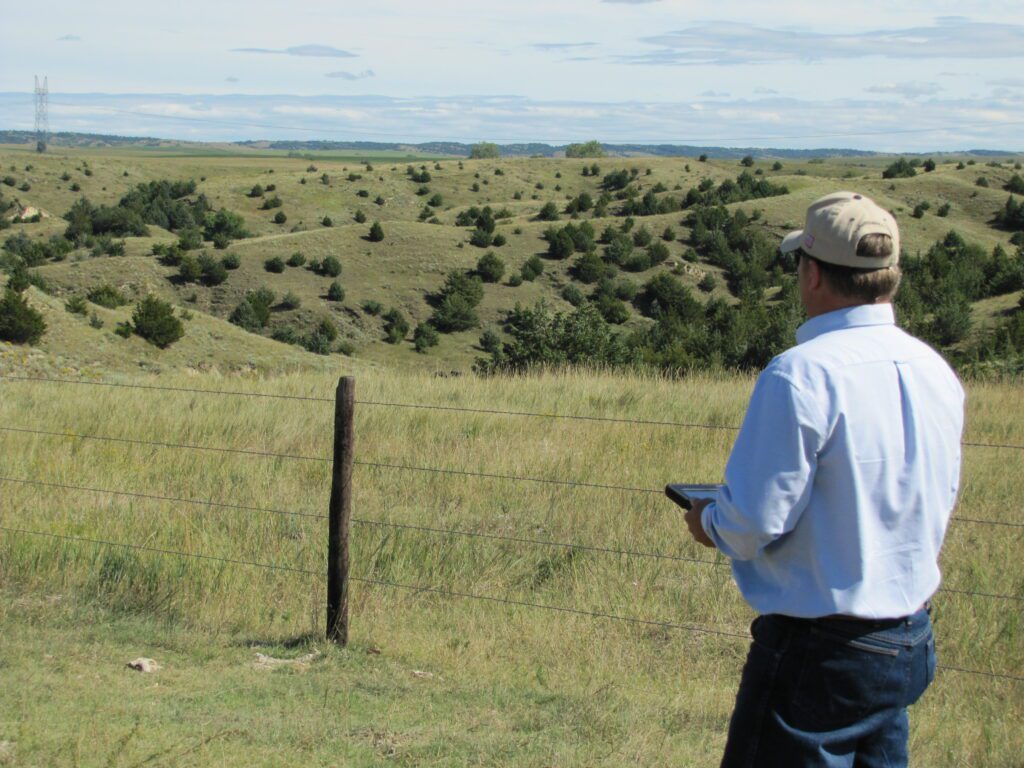 An image representing real estate appraisal services from Agri Affiliates. An Agri Affiliate appraiser overlooking a valley of Nebraska pasture land ripe with grass and trees.
